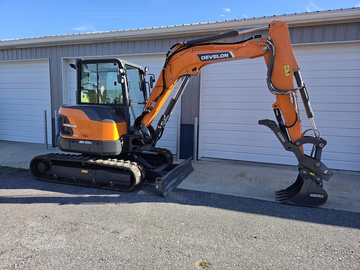 An orange Develon DX55R mini excavator parked in front of a metal storage building.