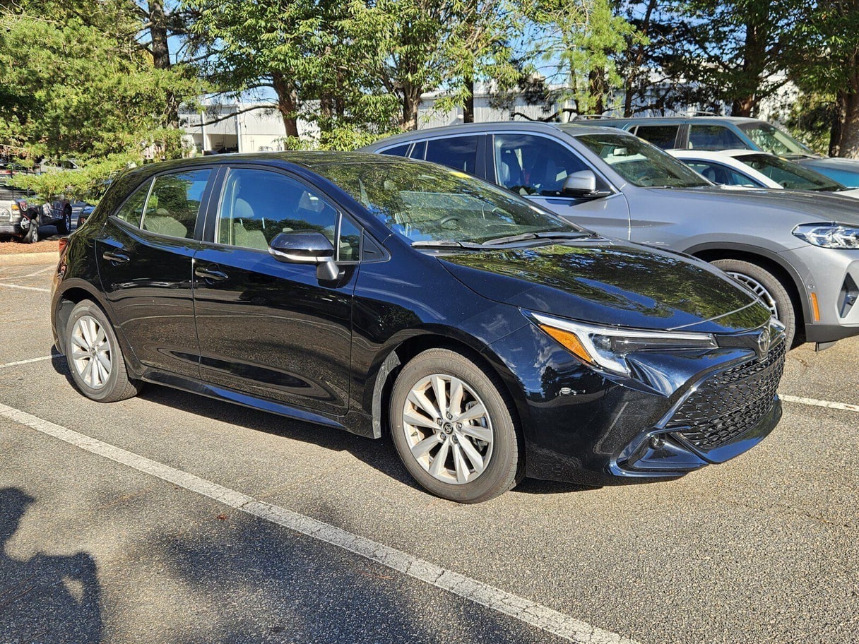 A black 2025 Toyota Corolla Hatchback parked beside other cars with trees behind it.
